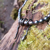 Oxidised silver spiral tendril pendant on black oval pearls, sitting on a mossy log in the sunshine