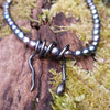 Oxidised silver spiral tendril pendant on black oval pearls, sitting on a mossy log in the sunshine