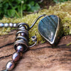 Oxidised black silver tendril pendant with blue flash labradorite bud winds around black oval freshwater pearls. Pictured on a mossy log. 