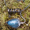 Oxidised black silver tendril pendant with blue flash labradorite bud winds around a chunky black silver spiga chain. Pictured on a mossy log