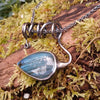 Oxidised black silver tendril pendant with blue flash labradorite bud winds around a chunky black silver spiga chain. Pictured on a mossy log