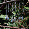 three pairs of oxidised silver dangly earrings with blue flash labradorite hanging from a twig with trees in the background.